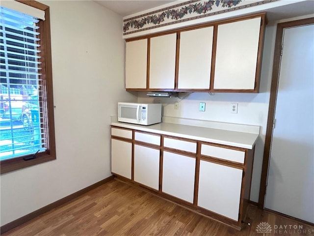 kitchen featuring wood finished floors, white microwave, baseboards, light countertops, and white cabinetry