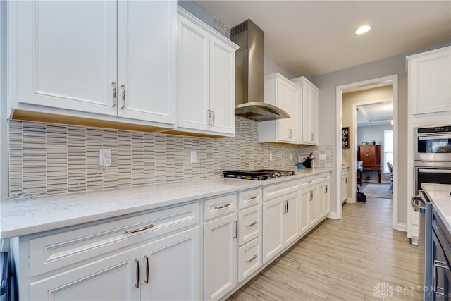 kitchen with light wood finished floors, appliances with stainless steel finishes, white cabinetry, wall chimney range hood, and backsplash