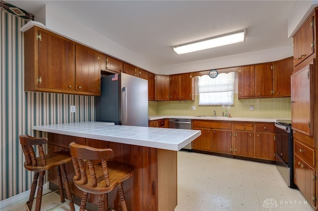 kitchen with brown cabinets, a sink, tile countertops, stainless steel appliances, and light floors