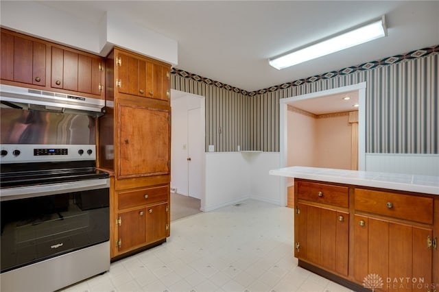 kitchen featuring brown cabinets, under cabinet range hood, wallpapered walls, stainless steel electric range oven, and light floors