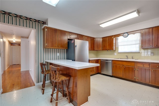 kitchen with brown cabinets, a sink, appliances with stainless steel finishes, wallpapered walls, and light floors