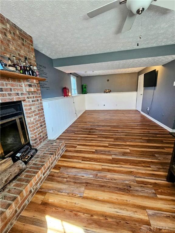 unfurnished living room featuring a ceiling fan, a textured ceiling, wood finished floors, a fireplace, and wainscoting