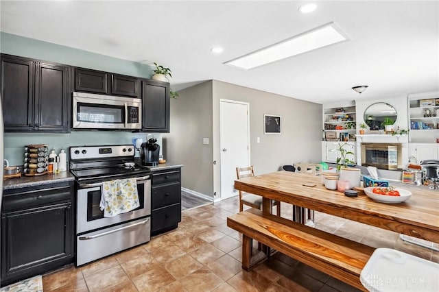 kitchen featuring a skylight, light tile patterned flooring, a glass covered fireplace, stainless steel appliances, and dark cabinets