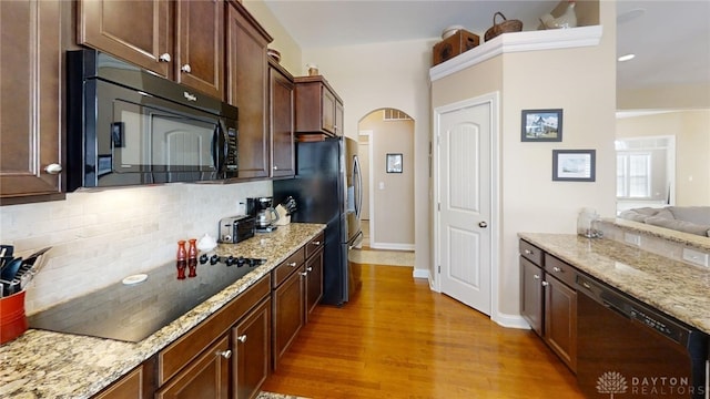 kitchen featuring light stone countertops, arched walkways, black appliances, light wood-style floors, and tasteful backsplash