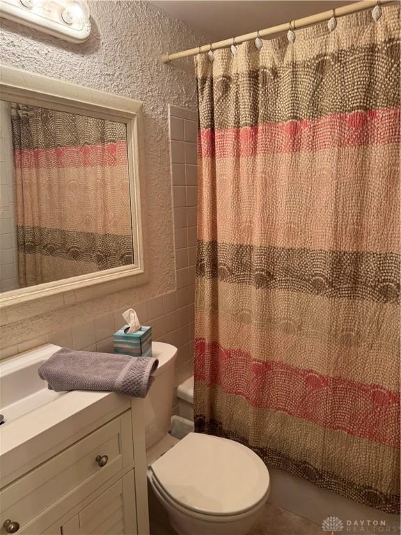 full bathroom with toilet, vanity, a textured wall, shower / bath combination with curtain, and tile walls