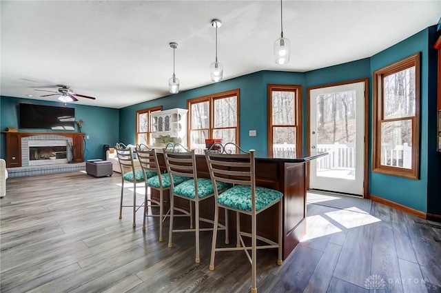 dining room featuring baseboards, a brick fireplace, wood finished floors, and a ceiling fan