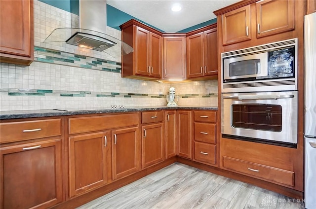 kitchen with dark stone countertops, brown cabinetry, appliances with stainless steel finishes, wall chimney range hood, and tasteful backsplash