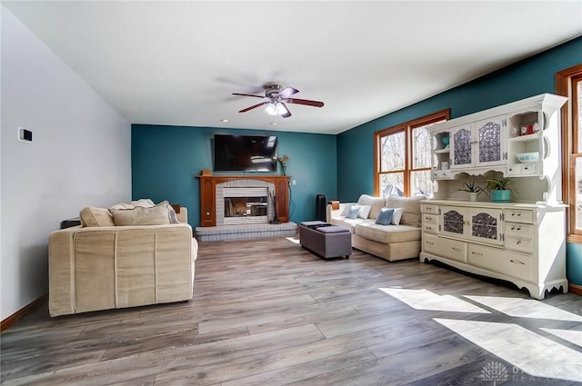 living room featuring a brick fireplace, wood finished floors, baseboards, and ceiling fan