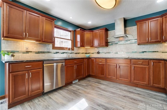 kitchen with wall chimney range hood, dark stone counters, stainless steel dishwasher, light wood-style floors, and a sink