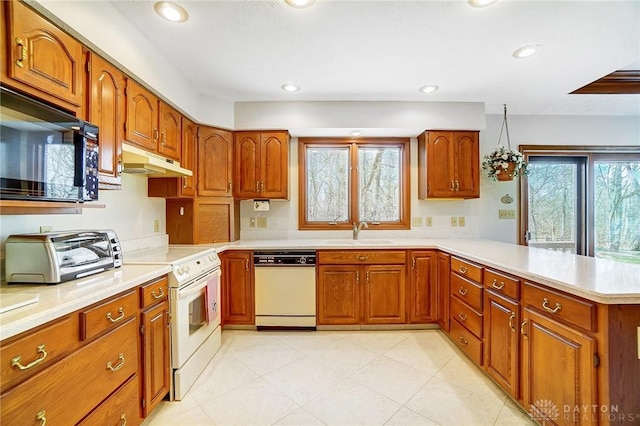 kitchen featuring brown cabinets, under cabinet range hood, a sink, white appliances, and a peninsula