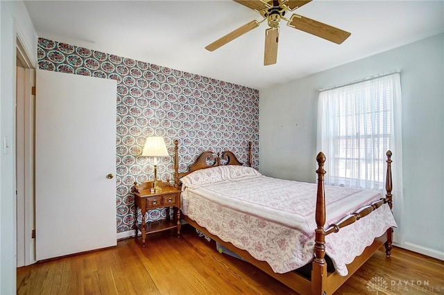 bedroom with wallpapered walls, an accent wall, ceiling fan, baseboards, and wood finished floors