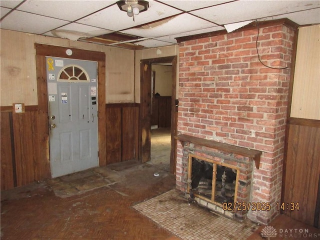 foyer featuring a paneled ceiling, wooden walls, a brick fireplace, and wainscoting