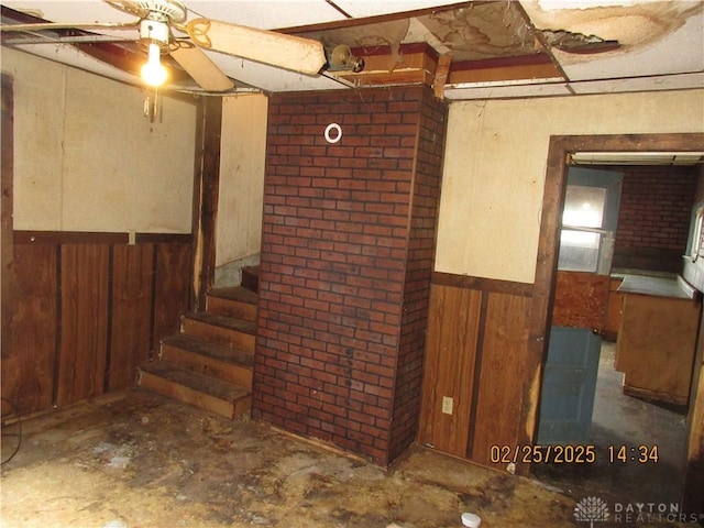 below grade area with stairway, a ceiling fan, a wainscoted wall, brick wall, and wood walls