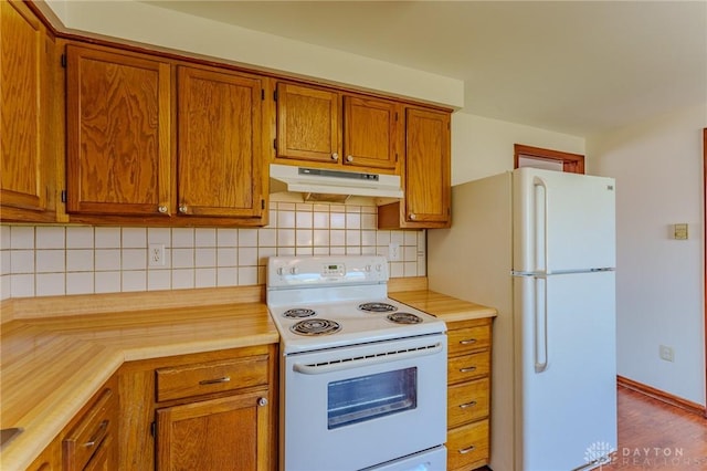 kitchen featuring white appliances, brown cabinetry, light countertops, under cabinet range hood, and backsplash