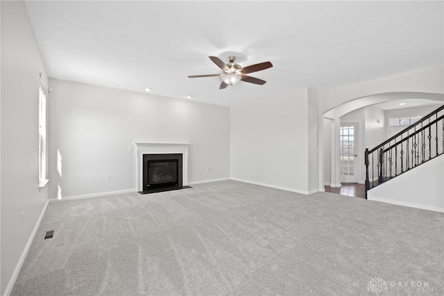 unfurnished living room featuring a fireplace with flush hearth, arched walkways, carpet, baseboards, and ceiling fan