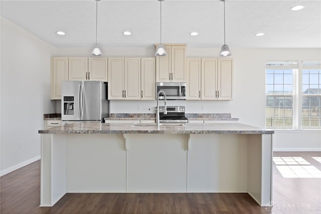 kitchen featuring dark wood-style floors, baseboards, an island with sink, cream cabinetry, and appliances with stainless steel finishes