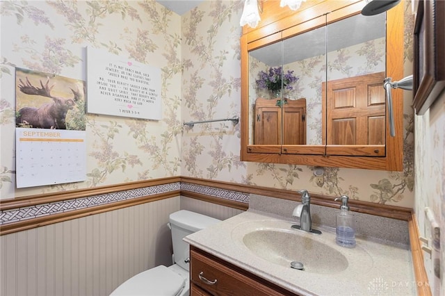 bathroom featuring vanity, a wainscoted wall, toilet, and wallpapered walls