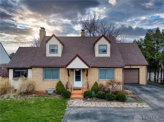 cape cod home with brick siding, aphalt driveway, an attached garage, and a chimney