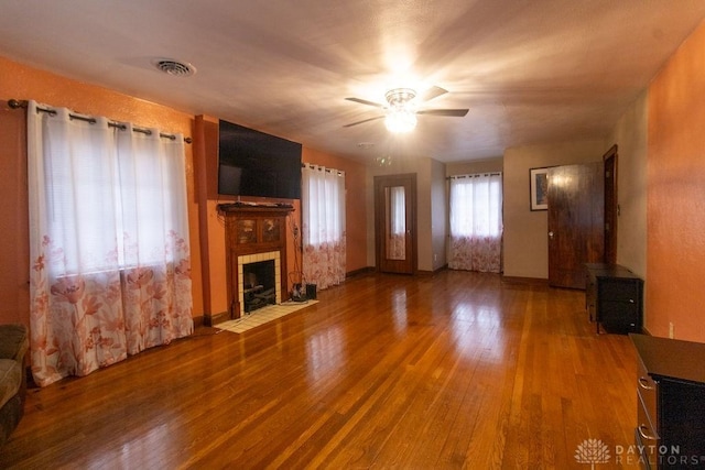 unfurnished living room featuring wood-type flooring, baseboards, visible vents, a tiled fireplace, and a ceiling fan