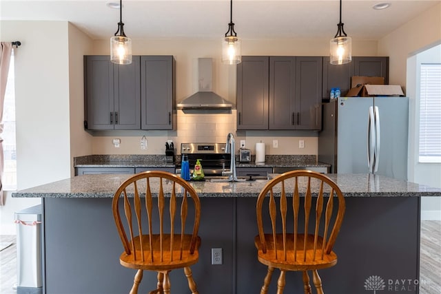 kitchen with dark stone countertops, wall chimney range hood, a center island with sink, a breakfast bar area, and stainless steel appliances