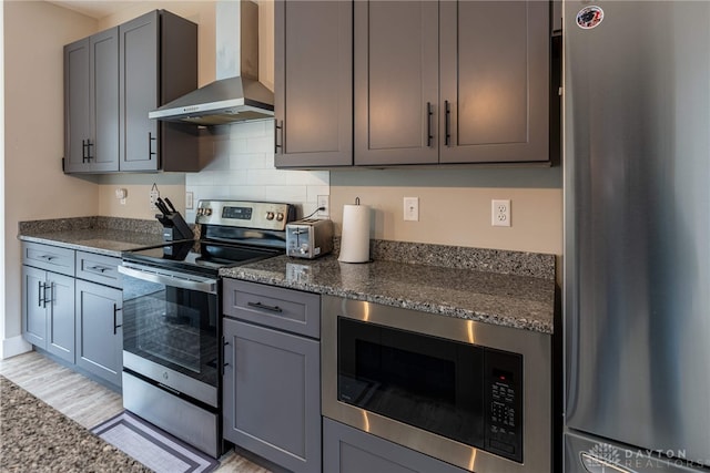 kitchen with dark stone counters, wall chimney exhaust hood, gray cabinets, appliances with stainless steel finishes, and backsplash