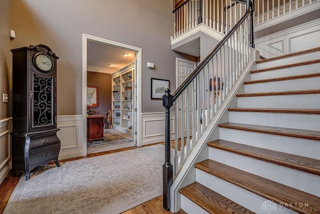 staircase featuring a decorative wall, crown molding, wainscoting, a towering ceiling, and wood finished floors
