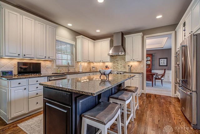 kitchen featuring wall chimney range hood, white cabinetry, stainless steel appliances, a kitchen island, and a breakfast bar area