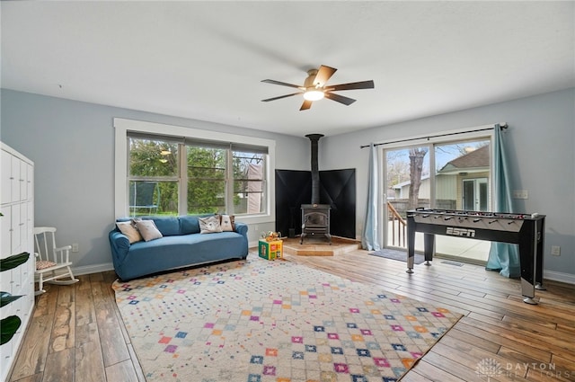 living area featuring wood-type flooring, a wood stove, a ceiling fan, and baseboards
