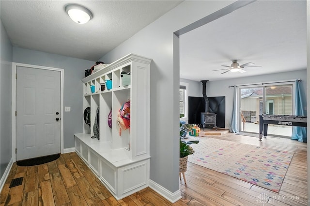 mudroom with a ceiling fan, a textured ceiling, a wood stove, wood-type flooring, and baseboards