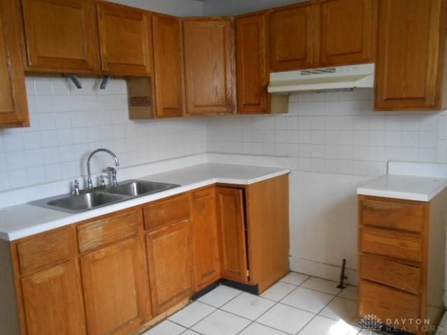 kitchen featuring brown cabinets, under cabinet range hood, light countertops, a sink, and tasteful backsplash