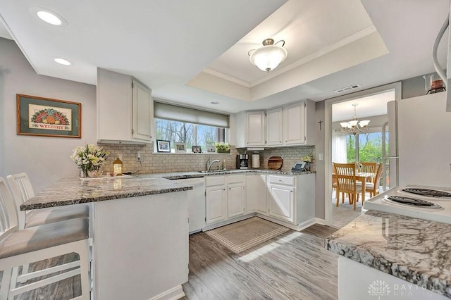 kitchen with light wood finished floors, an inviting chandelier, a tray ceiling, white appliances, and a peninsula