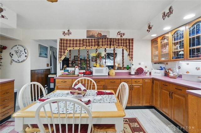 kitchen with brown cabinetry, light wood-style floors, glass insert cabinets, white microwave, and backsplash