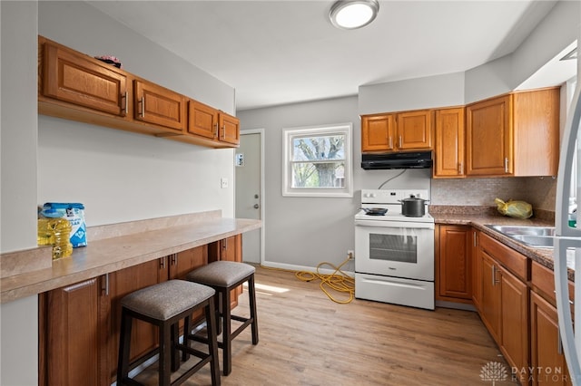 kitchen with light wood finished floors, a kitchen breakfast bar, brown cabinets, under cabinet range hood, and white range with electric cooktop