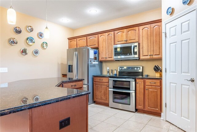 kitchen with brown cabinetry, dark stone counters, appliances with stainless steel finishes, light tile patterned floors, and decorative light fixtures