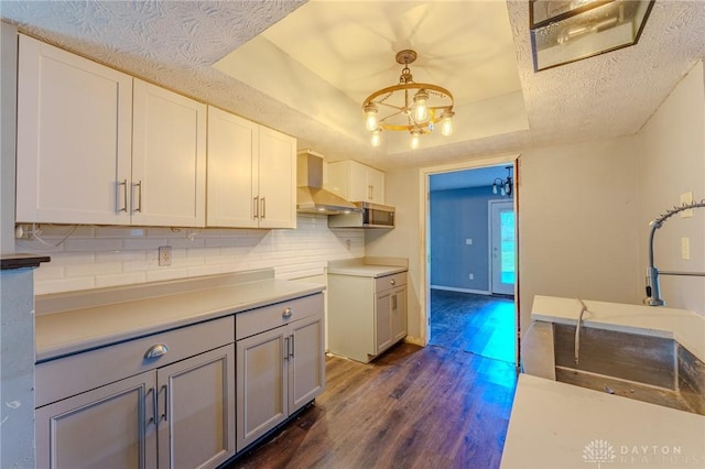 kitchen with wall chimney range hood, a tray ceiling, an inviting chandelier, dark wood finished floors, and backsplash