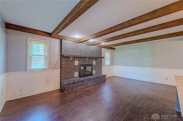 unfurnished living room with beam ceiling, wood finished floors, a wainscoted wall, and a brick fireplace