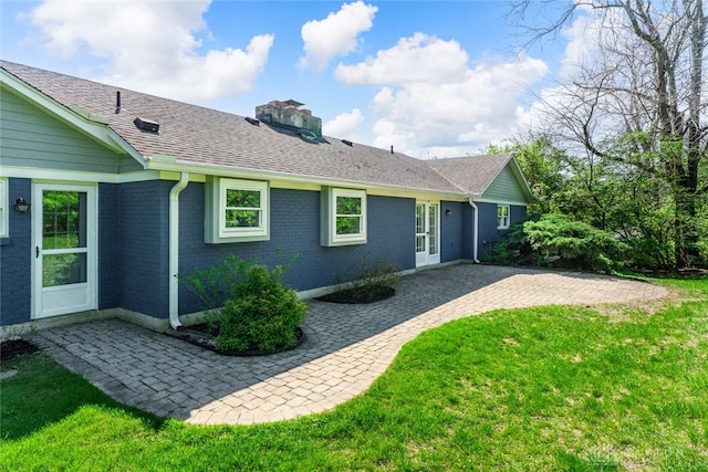 back of house featuring a chimney, brick siding, a shingled roof, a patio area, and a lawn