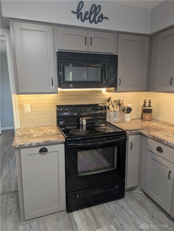 kitchen featuring gray cabinets, light stone countertops, decorative backsplash, black appliances, and light wood-type flooring