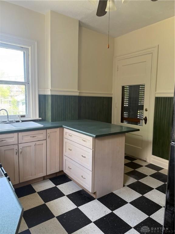 kitchen with light floors, a wainscoted wall, ceiling fan, a sink, and a peninsula