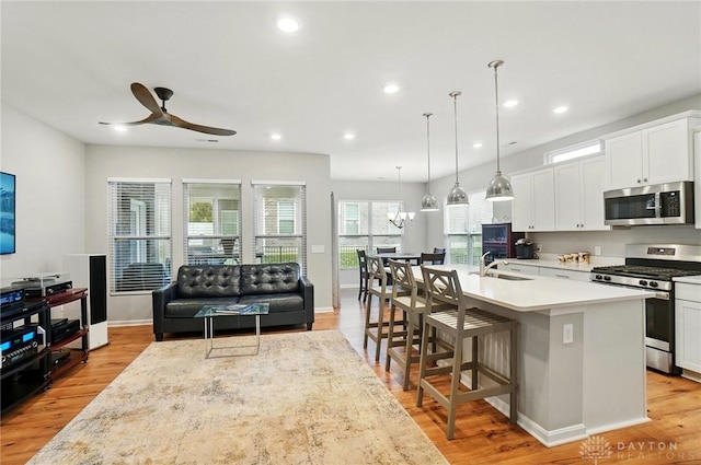 kitchen featuring an island with sink, light countertops, a sink, ceiling fan with notable chandelier, and stainless steel appliances