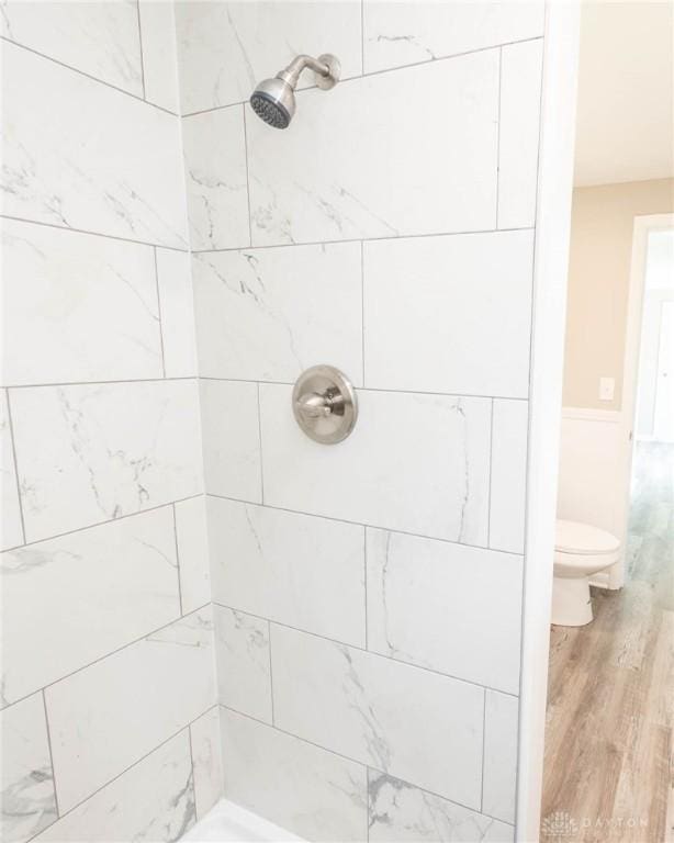 bathroom featuring wood finished floors, a tile shower, toilet, and a wainscoted wall