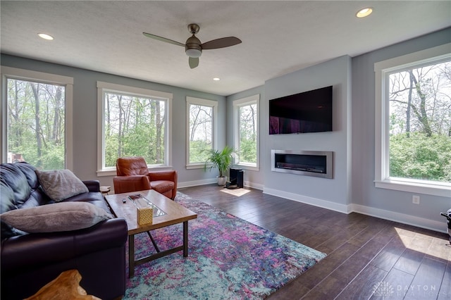 living area featuring a glass covered fireplace, baseboards, ceiling fan, and dark wood-style flooring