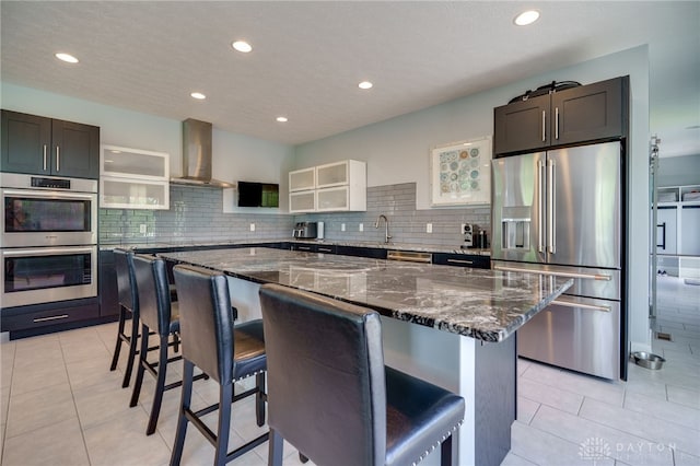kitchen with wall chimney range hood, stainless steel appliances, tasteful backsplash, and dark stone countertops
