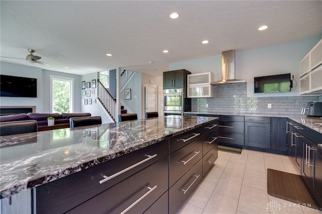 kitchen featuring wall chimney range hood, dark stone counters, a ceiling fan, and recessed lighting