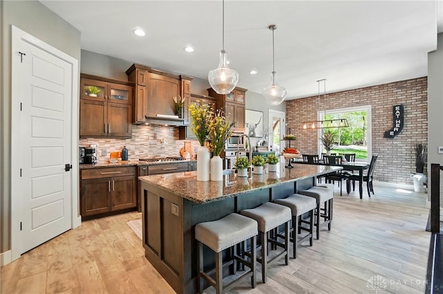 kitchen with dark stone countertops, light wood finished floors, brick wall, and an island with sink