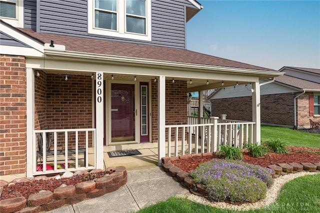 entrance to property featuring covered porch, a shingled roof, and brick siding