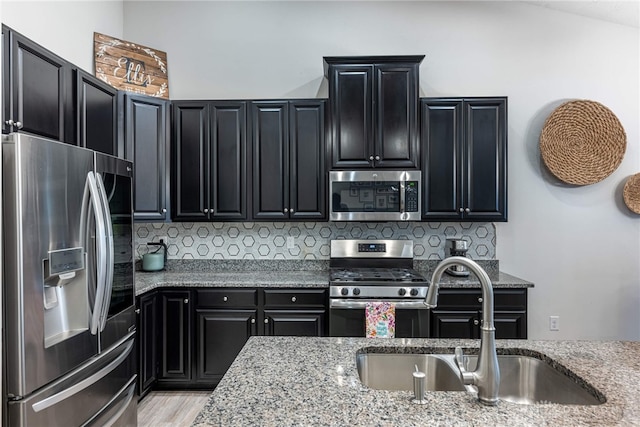 kitchen with backsplash, appliances with stainless steel finishes, light stone countertops, a sink, and dark cabinets