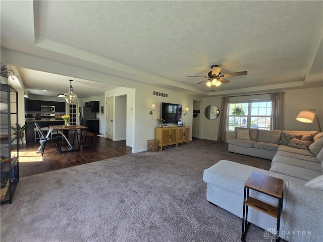 living room featuring ceiling fan, dark carpet, baseboards, a tray ceiling, and visible vents