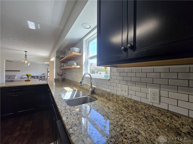 kitchen with open shelves, dark cabinets, light stone counters, a sink, and backsplash