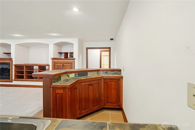kitchen featuring a fireplace, brown cabinetry, tile countertops, baseboards, and a peninsula